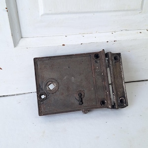 May include: An antique, rectangular, metal door lock with a keyhole and a square opening. The lock is a dark, rusty brown color and has a hinged plate attached to the side. The background is a white surface.