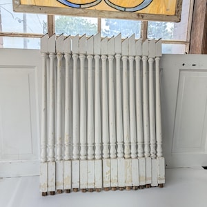 May include: A set of fifteen white painted wooden balusters arranged in a row. Each baluster features a turned spindle design with a square base and a slanted top. The white paint shows signs of wear and chipping, revealing the wood underneath.