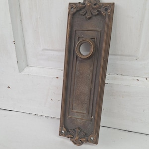 May include: An antique bronze door plate with ornate detailing. The rectangular plate features a circular opening and decorative flourishes at the top and bottom. The plate is set against a white, weathered wooden surface.