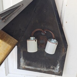 May include: A dark metal wall-mounted light fixture with a triangular backplate. The interior features two white ceramic light bulb sockets connected by brown electrical wiring. The fixture has a hinged metal panel and a decorative finial at the top.