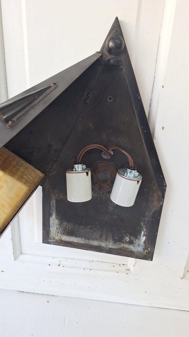 May include: A dark metal wall-mounted light fixture with a triangular backplate. The interior features two white ceramic light bulb sockets connected by brown electrical wiring. The fixture has a hinged metal panel and a decorative finial at the top.