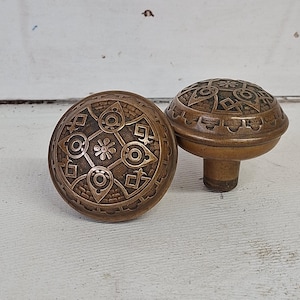 May include: Two antique bronze door knobs with intricate geometric patterns. One knob is lying on its side, while the other stands upright. The knobs have a warm, aged patina, suggesting they are vintage or vintage-inspired.