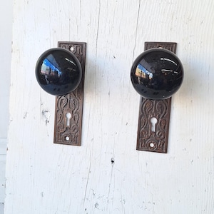 May include: Two antique door knobs with black, glossy spherical handles. The knobs are mounted on rectangular, ornate, bronze-colored backplates. The background is a white, weathered wooden surface.
