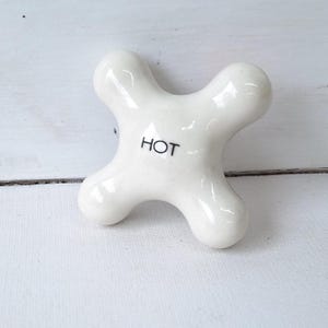May include: A white, four-pronged faucet handle with the word "HOT" printed in black. The handle has a glossy finish and is set against a white, textured background. This bathroom fixture is designed for controlling water temperature.