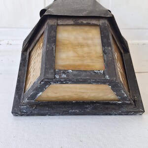 May include: A pyramid-shaped wall lantern featuring a dark, weathered metal frame with visible signs of wear and oxidation. The fixture includes translucent, amber-toned glass panels with a subtle marbled texture, set within the metal structure.