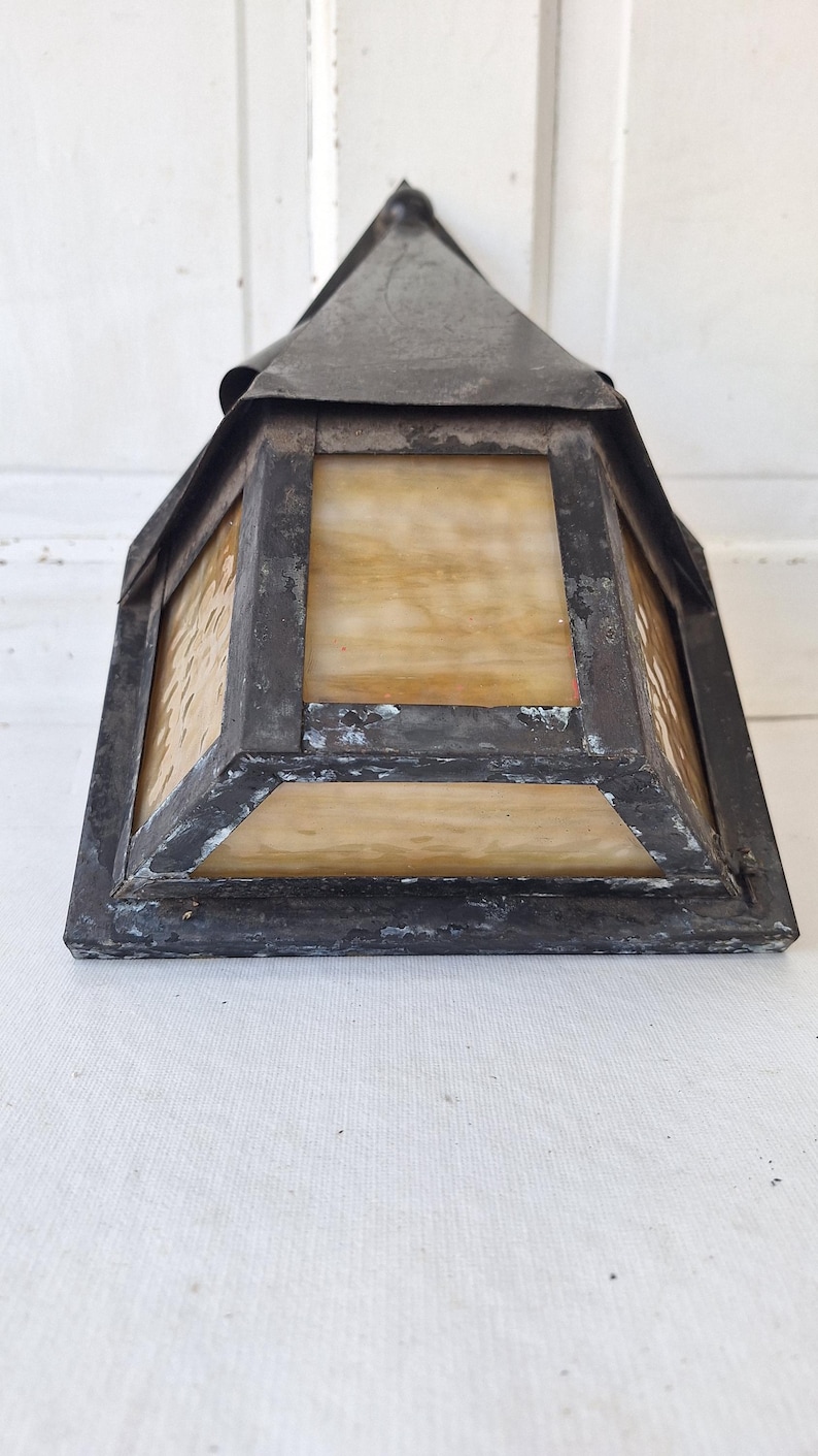 May include: A pyramid-shaped wall lantern featuring a dark, weathered metal frame with visible signs of wear and oxidation. The fixture includes translucent, amber-toned glass panels with a subtle marbled texture, set within the metal structure.
