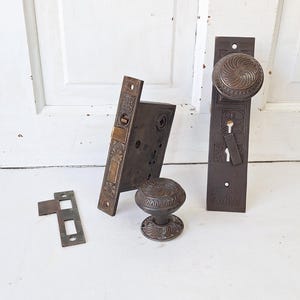 May include: Antique bronze door hardware set, including a doorknob, faceplate, and latch. The set features ornate, embossed detailing and a vintage aesthetic. The hardware is likely from the early 20th century.