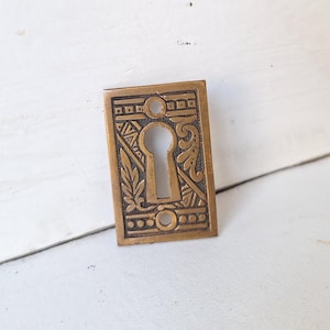 May include: A brass keyhole cover with an ornate design. The keyhole is surrounded by a decorative border with geometric and floral motifs.