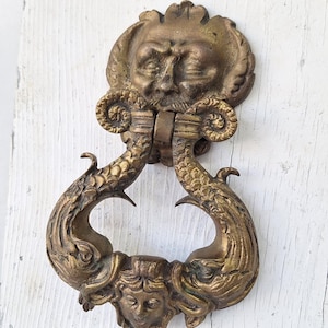 May include: Antique brass door knocker featuring a detailed design. The knocker has a face at the top and a mermaid-like figure at the bottom. The handle is shaped like two fish. The knocker is mounted on a white wooden door.