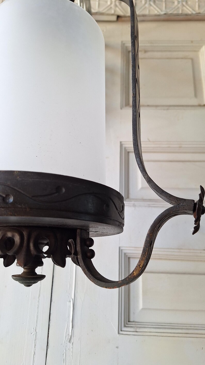 K&ouml;nnte beinhalten: Antiker Kronleuchter mit zylindrischem Schirm aus Milchglas und aufwendigen, dunklen Metallarbeiten. Das Metall weist dekorative Schn&ouml;rkel und einen gebogenen Arm auf. Die Leuchte ist f&uuml;r die Deckenbeleuchtung konzipiert.