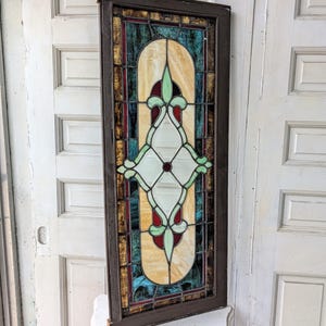 May include: A rectangular stained glass window panel encased in a dark brown wooden frame. The glass features a central vertical design with cream, light green, and deep red segments, surrounded by textured blue and gold glass border panels.