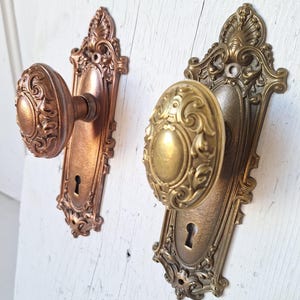 Puede incluir: Dos pomos de puerta antiguos con detalles ornamentados. Uno es de color cobre, el otro de lat&oacute;n. Ambos tienen cerraduras y placas decorativas. Los pomos est&aacute;n montados sobre una superficie de madera blanca envejecida.