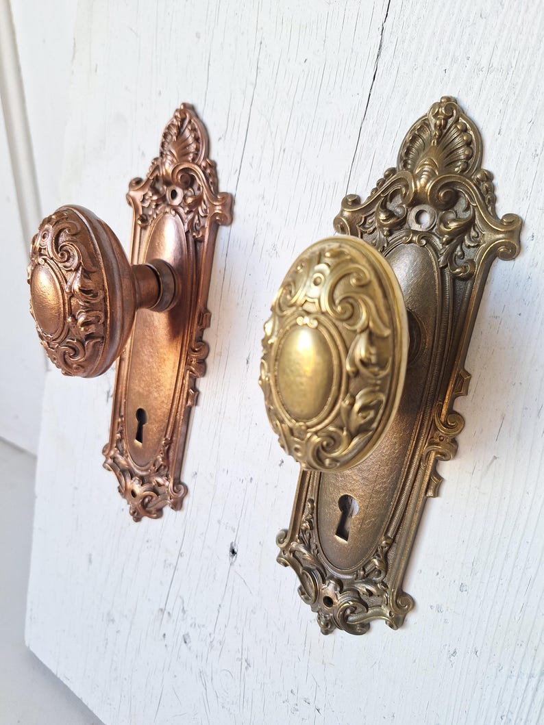 Puede incluir: Dos pomos de puerta antiguos con detalles ornamentados. Uno es de color cobre, el otro de lat&oacute;n. Ambos tienen cerraduras y placas decorativas. Los pomos est&aacute;n montados sobre una superficie de madera blanca envejecida.