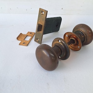 May include: A set of antique brass door hardware including two round knobs connected by a metal spindle, a rectangular latch plate with a dark textured body, and a small brass strike plate. The metal components show a weathered, aged patina.