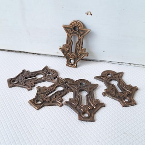 May include: A collection of antique bronze-colored keyhole covers. Each cover features an ornate, symmetrical design with a keyhole in the center and decorative details. The covers are arranged on a white surface.
