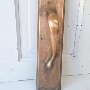 May include: A rectangular brass door handle plate with a curved, ergonomic grip. The metal surface features a weathered, aged patina with visible scuffs and dark oxidation marks. Four small circular screw holes are positioned at the corners.