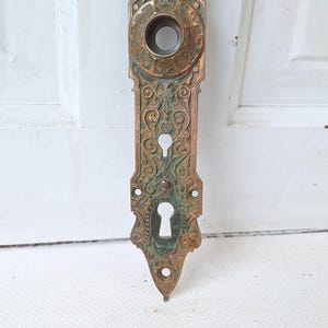 May include: Ornate antique door plate with a copper finish and green patina. The rectangular plate features a circular opening, two keyholes, and decorative scrollwork. A vintage hardware piece.