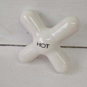 May include: A white ceramic faucet handle in a cross shape. The word "HOT" is printed in black on the center of the handle. The handle has a glossy finish and is likely for a bathroom or kitchen.