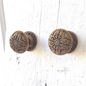May include: Two antique brass door knobs with intricate floral designs. The knobs are round and have a raised pattern of leaves and swirls.