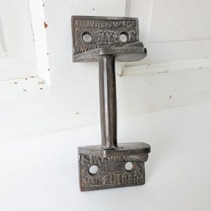 May include: A dark metallic door handle featuring two rectangular mounting plates with screw holes. The top plate is embossed with J.G. WILSON MFG CO N.Y. and the bottom plate displays PATENTED JAN. 21, 1890. The handle has a vertical cylindrical grip.