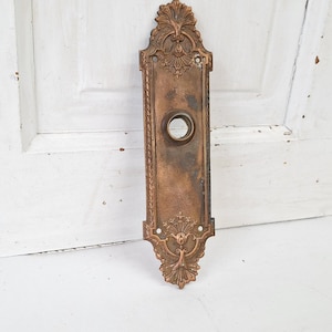 May include: Ornate antique bronze door plate with a central circular opening and decorative floral and scrollwork details. The rectangular plate has a weathered patina, suggesting age and use. The design is symmetrical, with embellishments at the top and bottom.