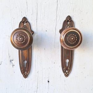 May include: Two antique bronze door knobs with matching backplates. The knobs are round with a floral design in the center, and the backplates have a keyhole and decorative details. The hardware is mounted on a white, weathered wooden surface.