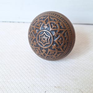 May include: A bronze-colored, round door knob with an intricate, embossed pattern. The design features a central six-pointed star surrounded by ornate, symmetrical shapes. The knob is likely made of metal and has a vintage or antique aesthetic.