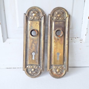 May include: A pair of rectangular brass door plates featuring an arched top and bottom with decorative embossed scrollwork. Each plate includes a central circular opening for a door knob and a lower keyhole opening, showing a weathered patina.