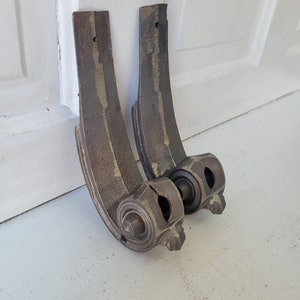 May include: Two antique metal scroll brackets with a decorative design. The brackets are a dark brown color with a gold patina. They are in good condition with some wear and tear.