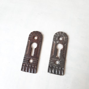 May include: Two antique keyhole escutcheons, one with a rusty finish and the other with a dark gray finish. Both have an ornate design and a keyhole in the center.