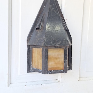 May include: A wall-mounted lantern featuring a dark, weathered metal frame with a pointed, triangular roof and translucent, amber-toned glass panels. The fixture has a hexagonal base and a distressed finish, showing signs of age and oxidation.