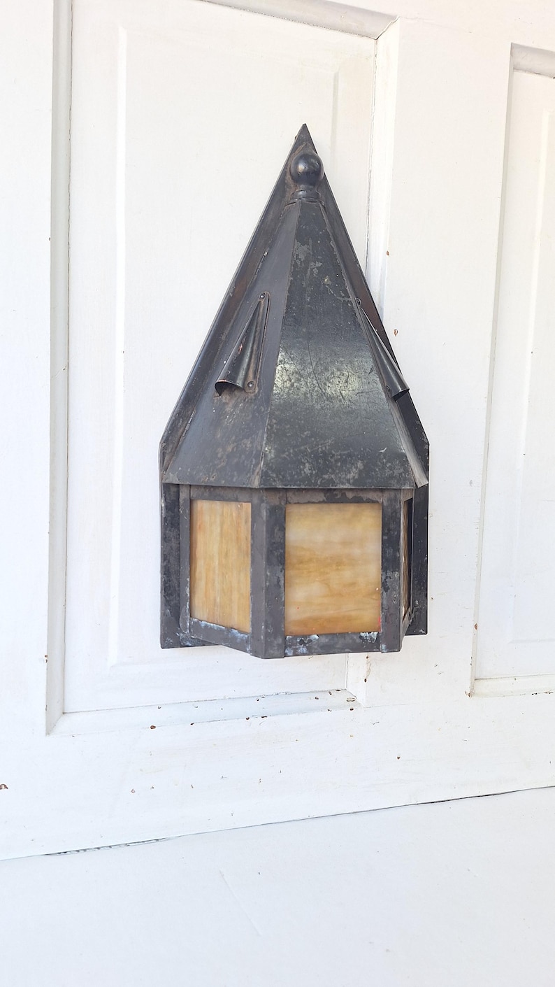 May include: A wall-mounted lantern featuring a dark, weathered metal frame with a pointed, triangular roof and translucent, amber-toned glass panels. The fixture has a hexagonal base and a distressed finish, showing signs of age and oxidation.