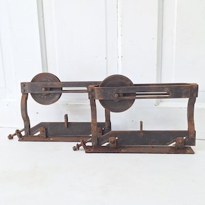 May include: Two antique, rusty metal pulley systems. Each system has a circular wheel, a rectangular base, and a series of rods and brackets. The systems are designed to be mounted on a wall or other surface. The background is a white door.