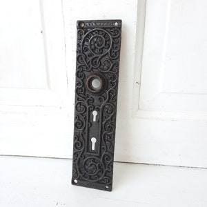 May include: Black cast iron door plate with an ornate design and two keyholes.