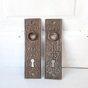May include: Two antique cast iron door plates with ornate designs and keyholes. The plates are a dark brown color with a rusty patina.