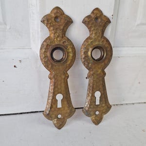 May include: Two antique brass door plates with a hammered texture. Each plate has a keyhole, a circular opening, and a decorative top. The plates are mounted on a white door.