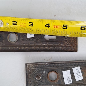 May include: Two antique, ornate bronze door plates with keyholes, each approximately 5 inches long, are shown with a yellow measuring tape. The plates have a detailed, embossed design and are likely vintage hardware.