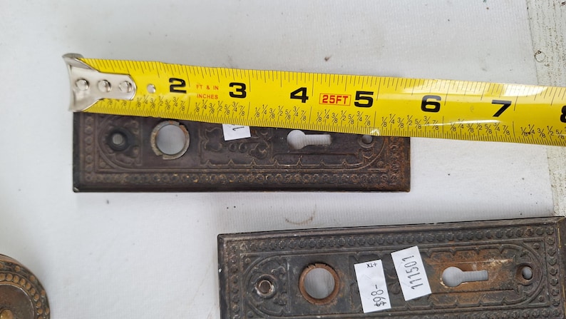 May include: Two antique, ornate bronze door plates with keyholes, each approximately 5 inches long, are shown with a yellow measuring tape. The plates have a detailed, embossed design and are likely vintage hardware.