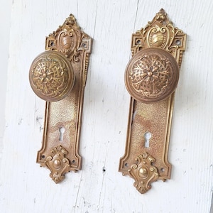 May include: Two antique brass door knobs with ornate designs and keyholes. The knobs have a textured surface and are mounted on rectangular plates with decorative edges.