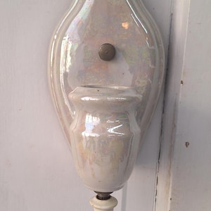 May include: A pearlescent white ceramic wall sconce with an oval backplate and a small vase-like holder. A small, round, metal button is centered on the backplate. The sconce has a small, white, cylindrical base. Home decor.
