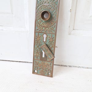 May include: An antique bronze door plate with intricate floral and geometric patterns. The rectangular plate features a keyhole, a circular knob, and a decorative key cover. The bronze has a green patina, adding to its vintage appeal.
