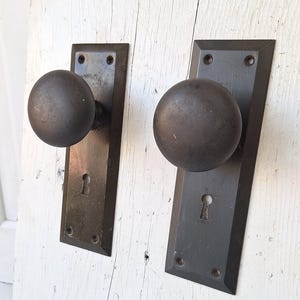 May include: Two antique door knobs with rectangular backplates. The knobs are round and dark gray, and the backplates have a keyhole. The door hardware is mounted on a white, weathered wooden surface.