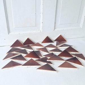 May include: A collection of brown triangular tiles with a glossy finish, arranged on a white surface. The tiles have a lighter brown hue on the edges and are set against a white door background. These tiles could be used for a mosaic or decorative project.