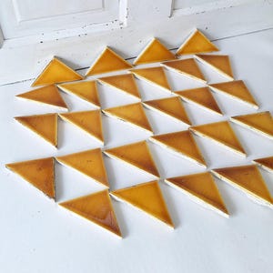 May include: A collection of triangular tiles arranged on a white surface. Each tile has a glossy, amber-colored top surface and a white base. The tiles are arranged in a grid-like pattern.