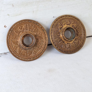 May include: Two antique bronze-colored door rosettes. Each rosette is circular with a central hole and intricate embossed floral and geometric patterns. The rosettes are displayed on a white surface.