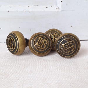 May include: A set of four antique brass door knobs. Each knob features a circular design with an embossed letter or monogram. The knobs have a dark, aged patina, suggesting they are vintage or reclaimed.