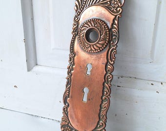 Pode incluir: Uma placa de porta retangular de metal em tom de cobre com um padrão em relevo ornamentado, motivos florais, uma abertura central circular para um puxador e duas ranhuras verticais para chave. A placa tem bordas arredondadas e orifícios.