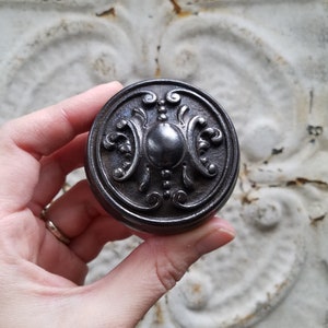 May include: A black metal cabinet knob with an ornate design. The knob has a raised center with a decorative pattern.