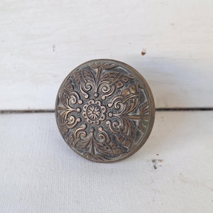 May include: Antique bronze-colored cabinet knob with an ornate floral design. The round knob features a central flower motif surrounded by swirling leaves and decorative patterns. The knob is likely made of metal and has a weathered appearance.
