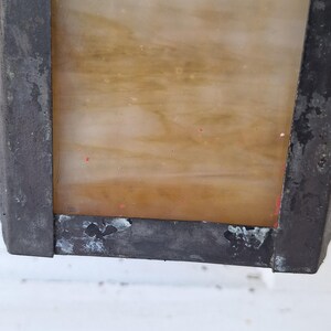 May include: A rectangular wall-mounted lantern featuring a weathered, dark metal frame with visible signs of oxidation and wear. The central panel consists of a translucent, amber-toned glass with subtle swirling patterns and small red paint flecks.